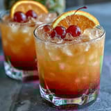 Beautiful layered sunrise mocktail featuring grenadine syrup, orange juice, and sparkling water, garnished with orange slices and cherries.